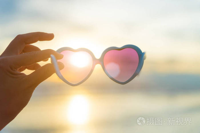 柔和的关注女性手拿太阳镜的心脏形状在海上和桑迪海滩的背景,在日落