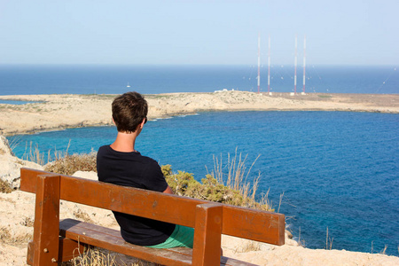阳光明媚的日子里,男人坐在长凳上,望着海洋和石岸.