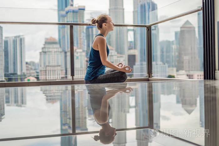 年轻的女人早上在她的阳台上练习瑜伽,可以看到城市和摩天大楼的全景.