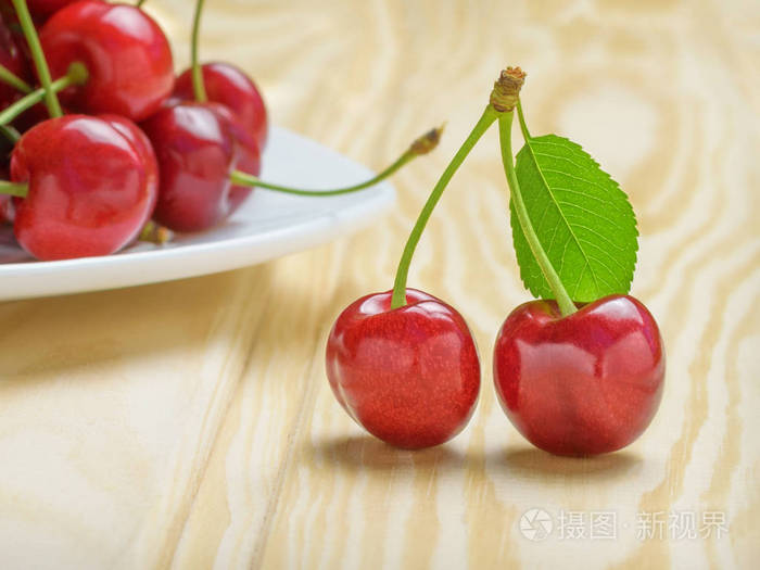 两个成熟的红色樱桃旁边是一片绿叶旁边是一个装满樱桃的白色盘子轻木