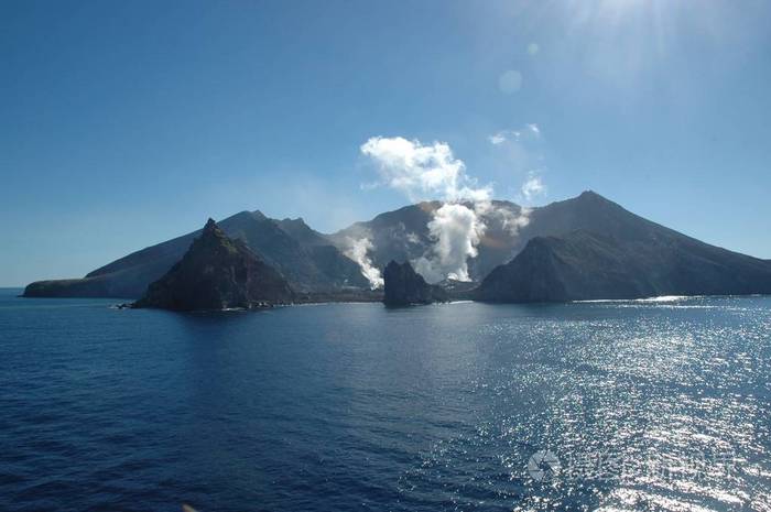 白烟和蒸汽从火山中升起。它在一个被蓝海包围的岛上。大海是平静的。天空是蓝色的，没有云。