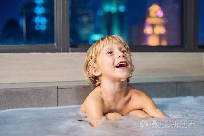 快乐的小男孩晚上坐在浴缸里，然后睡在窗户的背景下俯瞰傍晚的城市。 一幅婴儿在窗户附近满是泡沫的浴缸里洗澡的画像