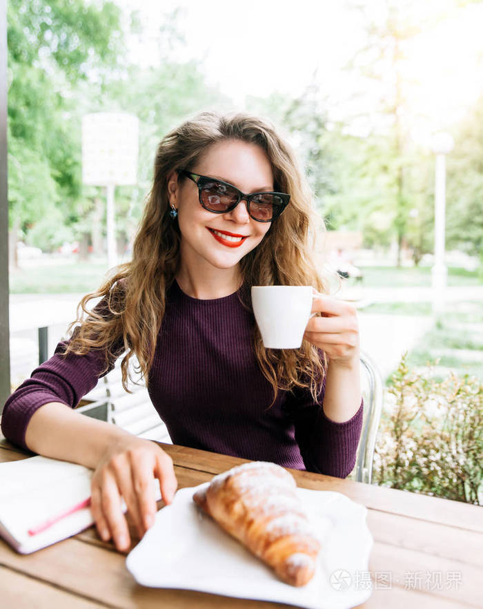 美丽迷人的年轻金发女人，卷发，喝卡布奇诺和吃牛角面包在城市咖啡馆夏天。 笔记本和电话在桌子上