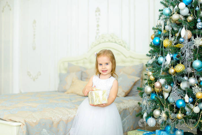 穿着白色连衣裙的小女孩在卧室装饰的圣诞树旁留着礼物