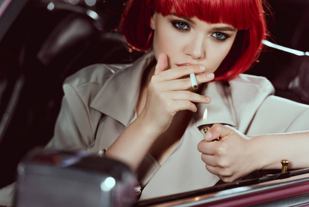 戴着红色假发的迷人女孩抽烟,坐在车里看着摄像机照片