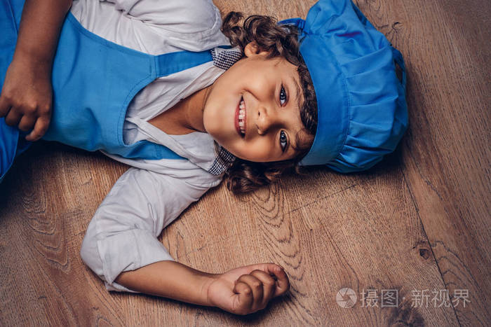 快乐的小男孩，一头棕色卷发，穿着蓝色的厨师制服，躺在木地板上