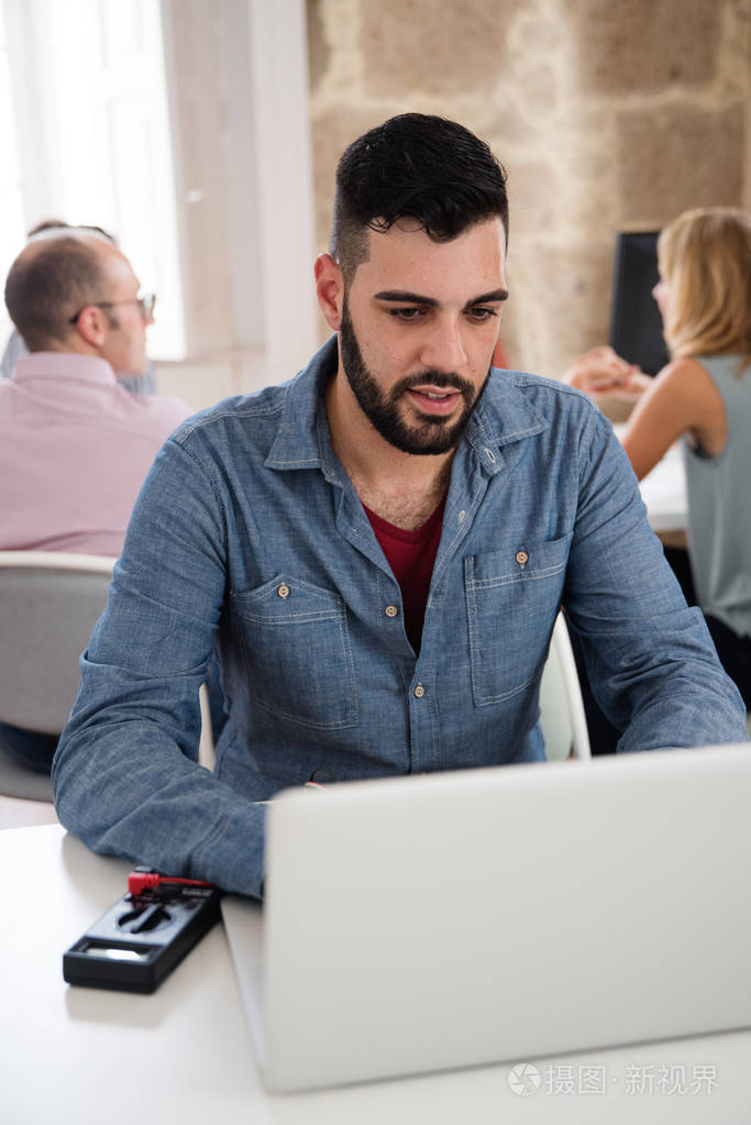 一个长胡子的男人,穿着蓝色衬衫,旁边有电器设备,坐在办公桌前,在