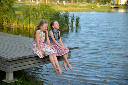 快乐的孩子们,小女孩,在日落时坐在河边的一座木桥上玩,光着脚聊天,笑
