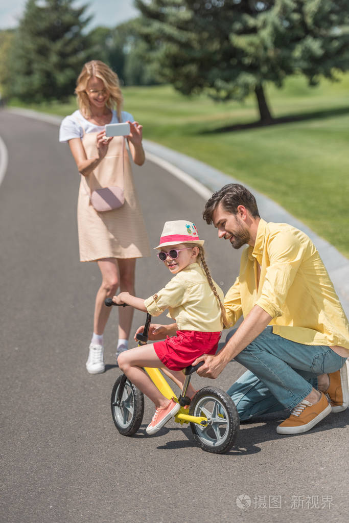 微笑的年轻女子带着智能手机拍照快乐的父亲教女儿骑自行车