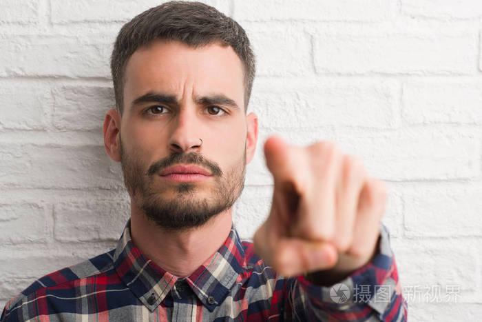年轻的成年人站在白色砖墙上用手指指着相机和你的手从前面签上积极和