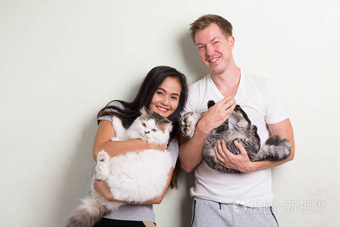 年轻快乐多族裔夫妇抱着两只猫在一起反对白色背景