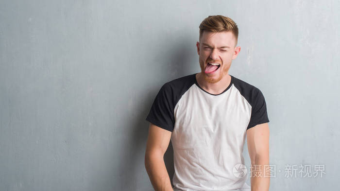年轻的红头发男人在灰色的墙壁上伸出舌头，以有趣的表情高兴。 情感概念。