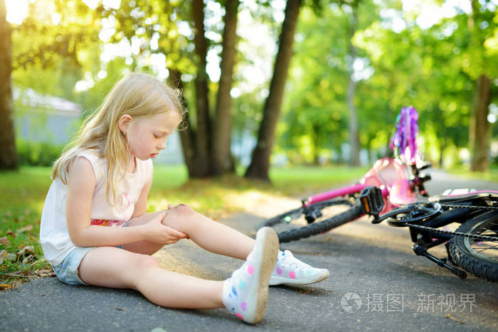 可爱的小女孩在夏天公园从自行车上摔下来后坐在地上。 孩子在骑自行车时受伤。 积极的家庭休闲与孩子。