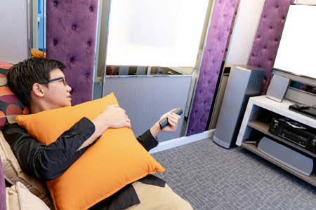 年轻的亚洲快乐男子使用遥控器,而看电视躺在沙发上的家庭影院房间.