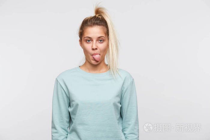 可爱的,金发和马尾辫的年轻女人的肖像,穿着蓝色的运动衫,露出舌头,在