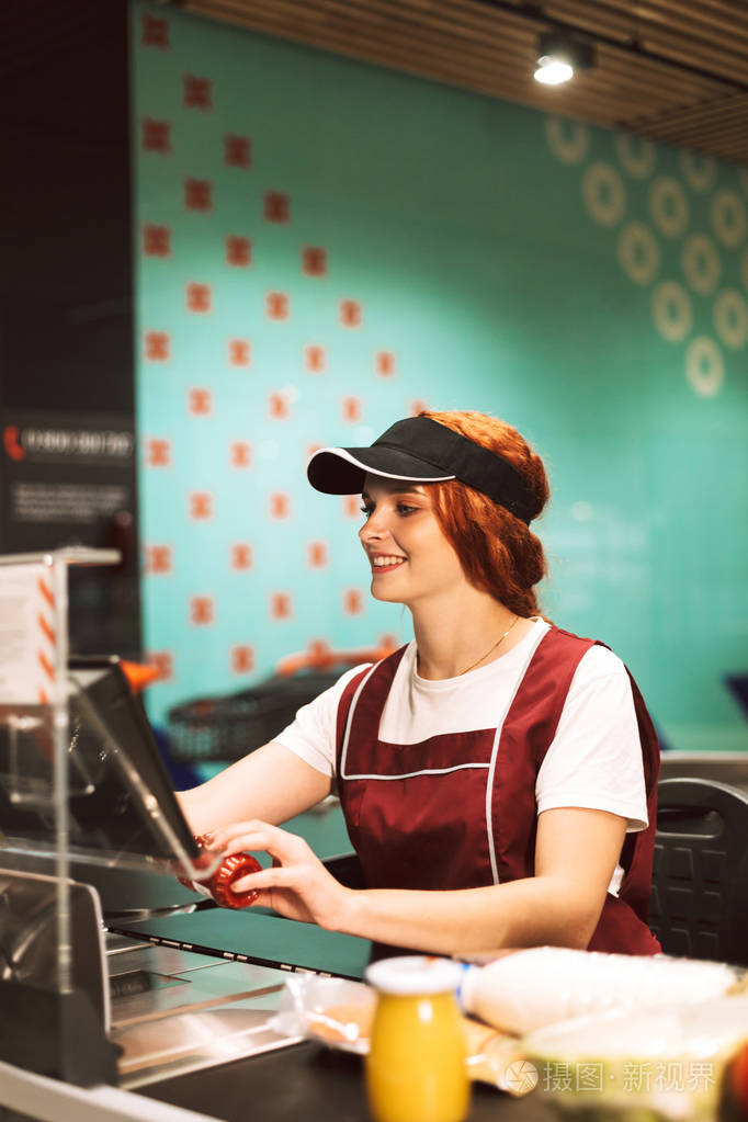 年轻微笑的女收银员穿着制服在现代超市工作愉快地使用现金箱