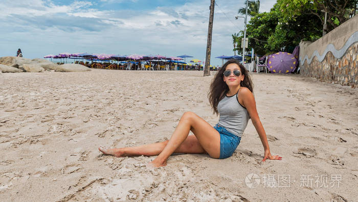 年轻女子戴着墨镜坐在沙滩上，背后撑着雨伞