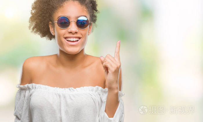 年轻的非洲美国女人戴着太阳镜，在孤立的背景上指着手指，提出了成功的想法。 退出并快乐。 一号。