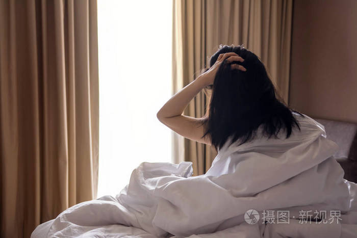 一个懒女人头发乱糟糟的坐在白床上早晨透过窗户看着外面