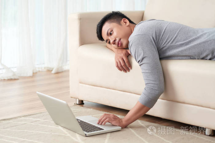 懒散的亚洲男人躺在沙发上看着笔记本电脑上的东西