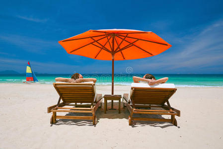 一对夫妇在热带海滩上，在红色雨伞下的甲板椅子上