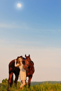 美丽的女孩在傍晚的田野里带着栗色的马
