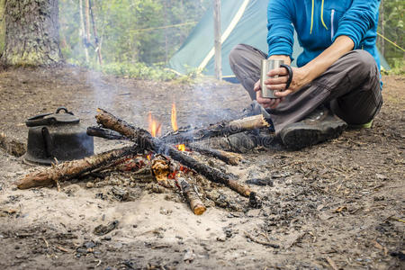 女孩坐在火旁露营时加热并喝热茶