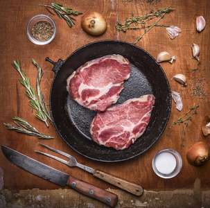 新鲜的生猪肉牛排，在铸铁煎锅上，用刀切肉叉，洋葱，大蒜，草药和香料。 木制乡村酒吧