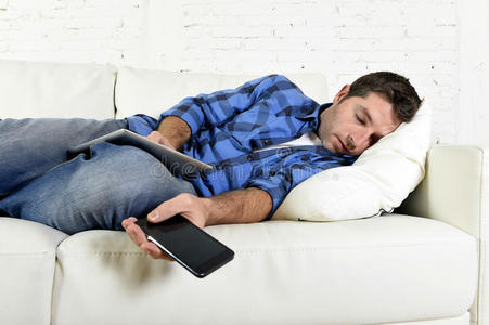 有吸引力的男人睡在家里的沙发上，手里拿着手机和数字平板电脑垫