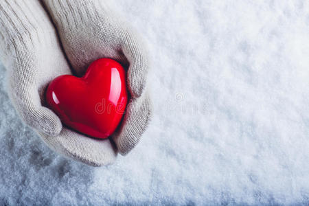 女性的手在白色针织手套与光泽红色的心在雪的背景。 爱和圣瓦伦丁的概念。
