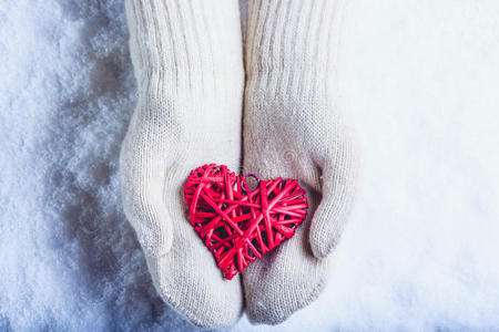 女性的手在白色针织手套与缠绕的复古浪漫红心在雪的背景。 爱和圣瓦伦丁的概念