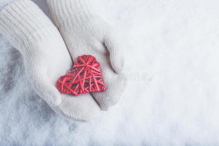 女性的手在白色针织手套与缠绕的复古浪漫红心在雪的背景。 爱和圣瓦伦丁的概念