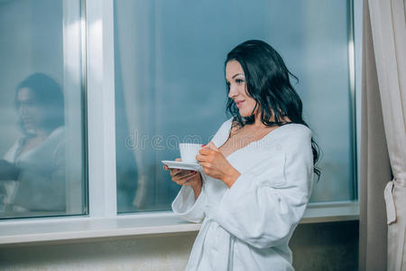 用新鲜咖啡取暖。 穿着白色浴袍的漂亮年轻女人喝着咖啡，透过窗户看着