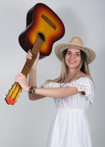 美丽的年轻的金发乡村女孩穿着一件白色的连衣裙和带吉他的牛仔帽