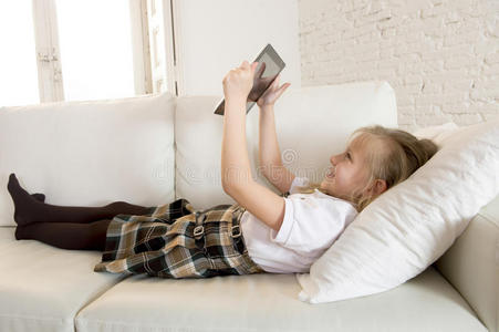 金发小女孩躺在家里的沙发上，在数字平板电脑垫上使用互联网应用程序