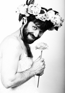 一个留着胡子的男人，穿着女人的婚纱，光着身子，手里拿着一朵花。他头上戴着花环。有趣的