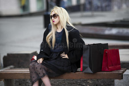 美丽的年轻时尚金发女人，穿着黑色外套和太阳镜，购物袋坐在公园的长凳上