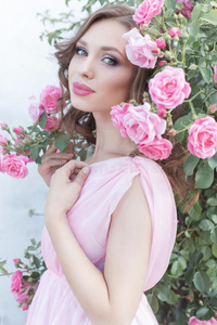美丽的女孩穿着粉红色的连衣裙，站在花园里的玫瑰，在阳光明媚的夏天，用温柔的化妆和明亮的PUF