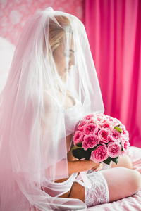 美丽的年轻新娘内衣坐在床上，手里拿着一束花，头上戴着面纱。 早上好