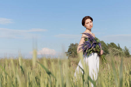 美丽的甜美女孩，留着辫子，穿着白色的夏季日裙，走在一片田野里，手里拿着一束紫色的卢平花