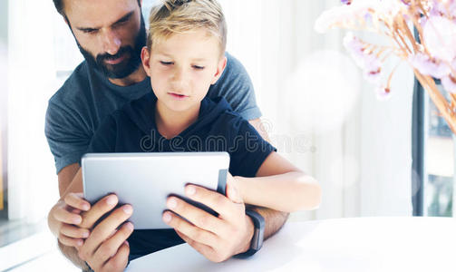 大胡子男人和小男孩在阳光下用平板电脑房间，爸爸和小儿子一起在移动电脑上玩耍，休息