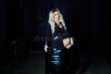 穿着黑色衣服的漂亮时尚金发女人坐在金属桶上。 完全是黑暗的风格