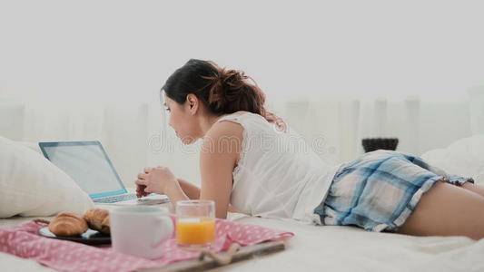 黑发女孩早上躺在床上聊天，用笔记本电脑。 年轻的女人在键盘上打字，笑着。