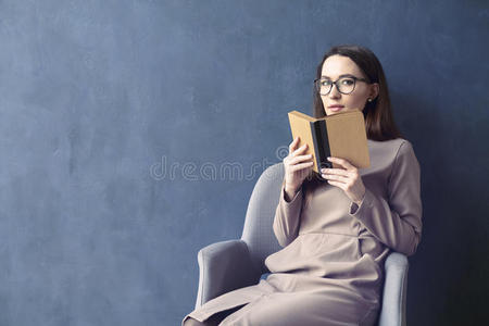 坐在阁楼办公室里读古董书的漂亮女商人。 打开书棕色封面。 深蓝色墙壁背景，白天光线