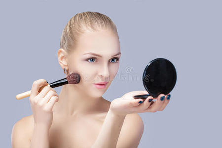 美丽的金发女人拿着化妆刷，在灰色背景上看着镜子。