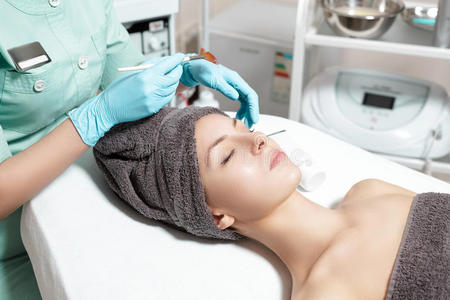 美容师在水疗沙龙中为美丽的年轻女性涂上带刷子的面膜。 化妆品程序皮肤护理。