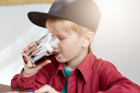 一个可爱的小男孩在夏天坐在餐馆里喝果汁时戴着时尚帽子的特写镜头。 那个拿着一杯的男孩