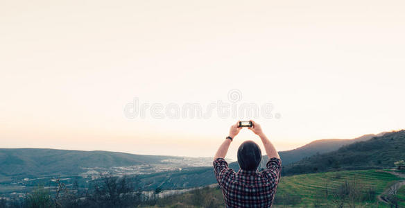 一个年轻人在日落时站在山顶上拍摄天空的照片