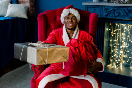 快乐微笑的非洲圣诞老人在他舒适的房子里，壁炉旁，圣诞树坐在扶手椅上，拿着礼物盒。圣诞节假期