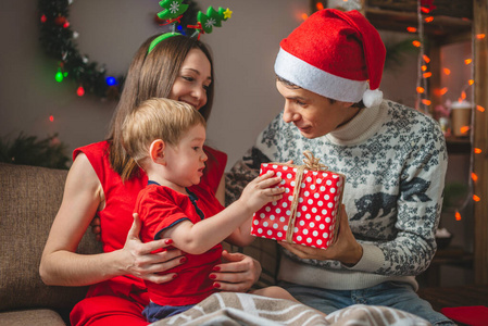 妈妈和爸爸给他们的儿子一个红色盒子礼物。带孩子的年轻家庭在家庆祝圣诞节和新年